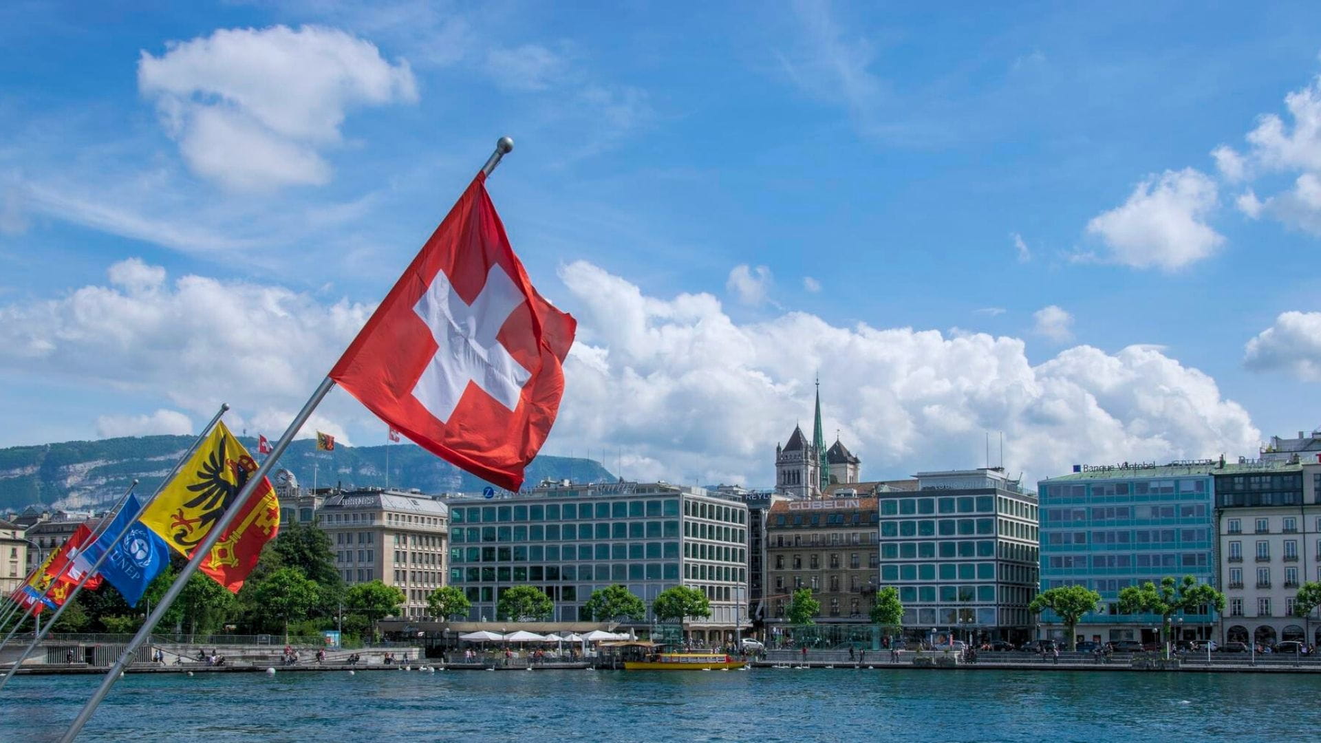 Swiss flag waving in Geneva with the cantonal flag and city buildings under a bright blue sky.